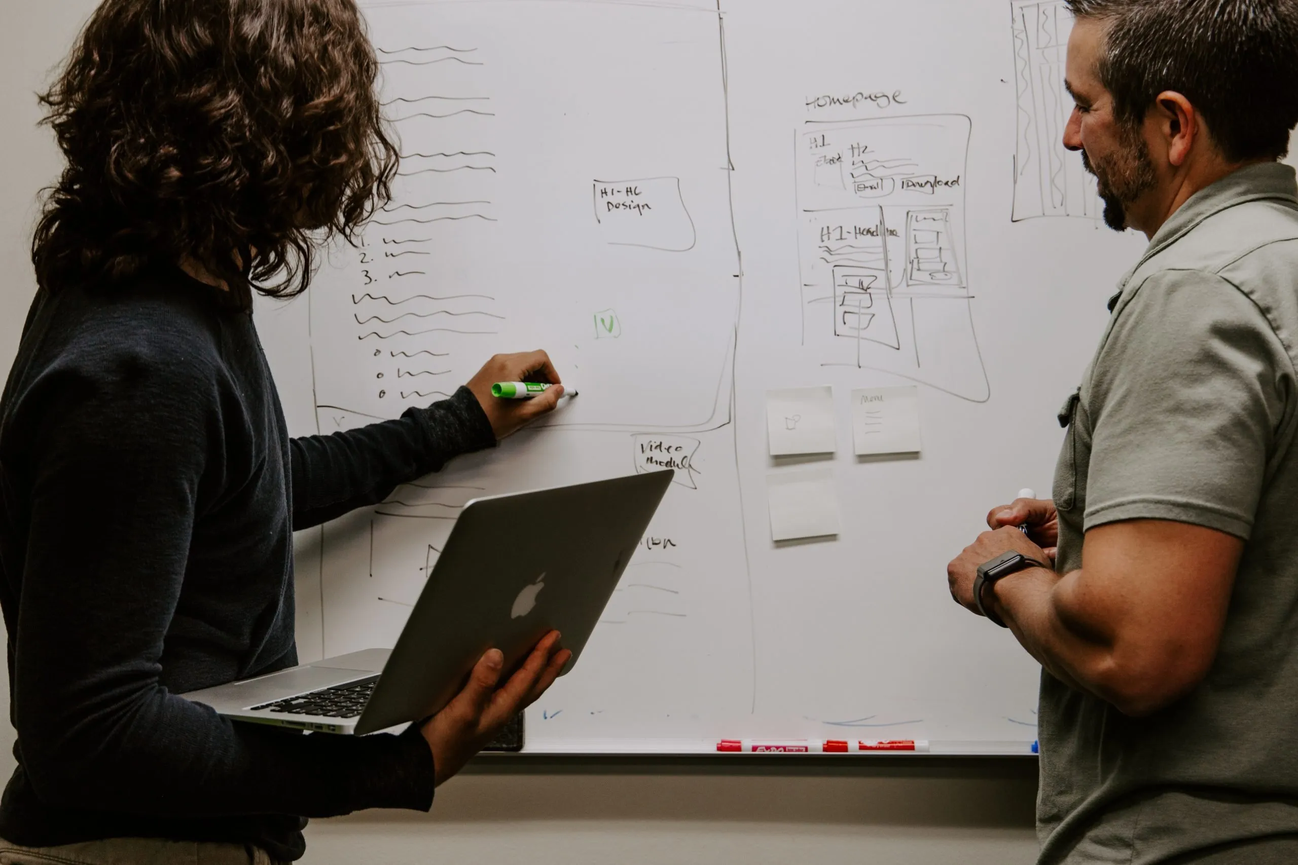 woman drawing on a whiteboard while holding a laptop in front of a colleague
