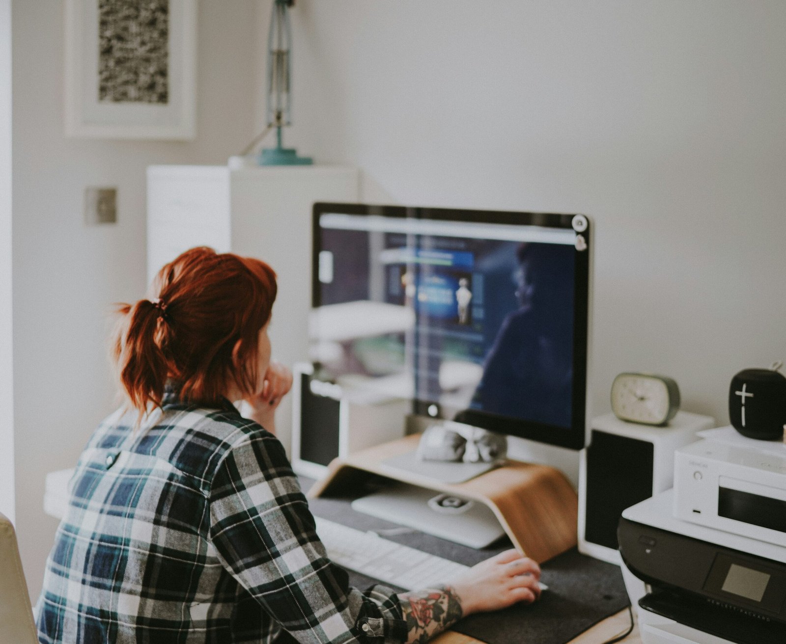 A woman sits at a desk in a home office, focused on a computer screen.