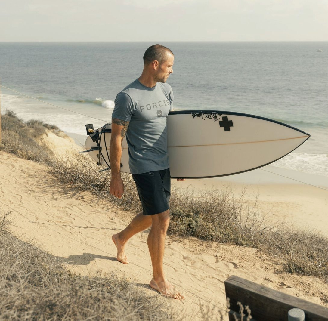 Man in a Forcis t-shirt carrying a surfboard down a beach path.