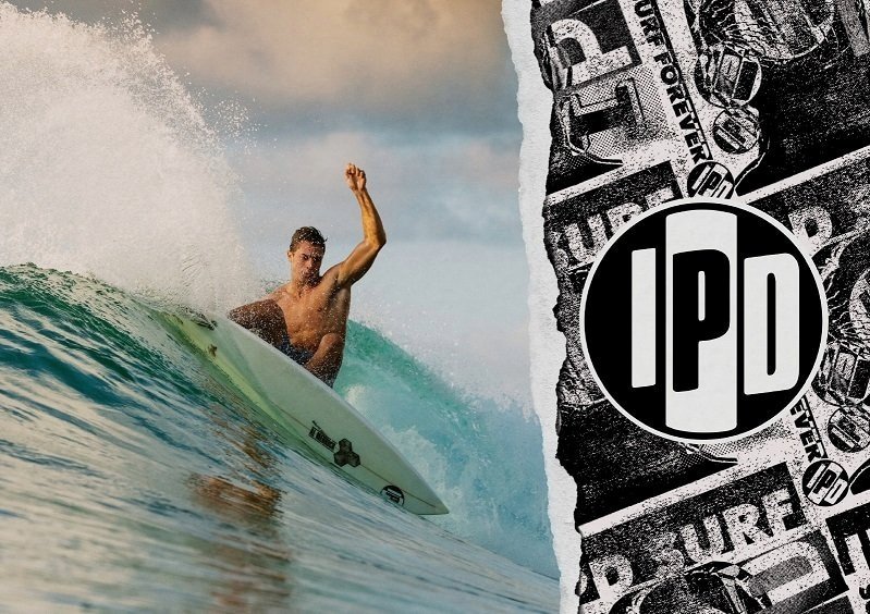 Surfer riding a wave with fist raised beside a torn I P D logo graphic.