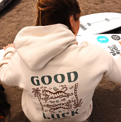 Cream 'Good Luck' graphic hoodie worn on a beach with a surfboard nearby.