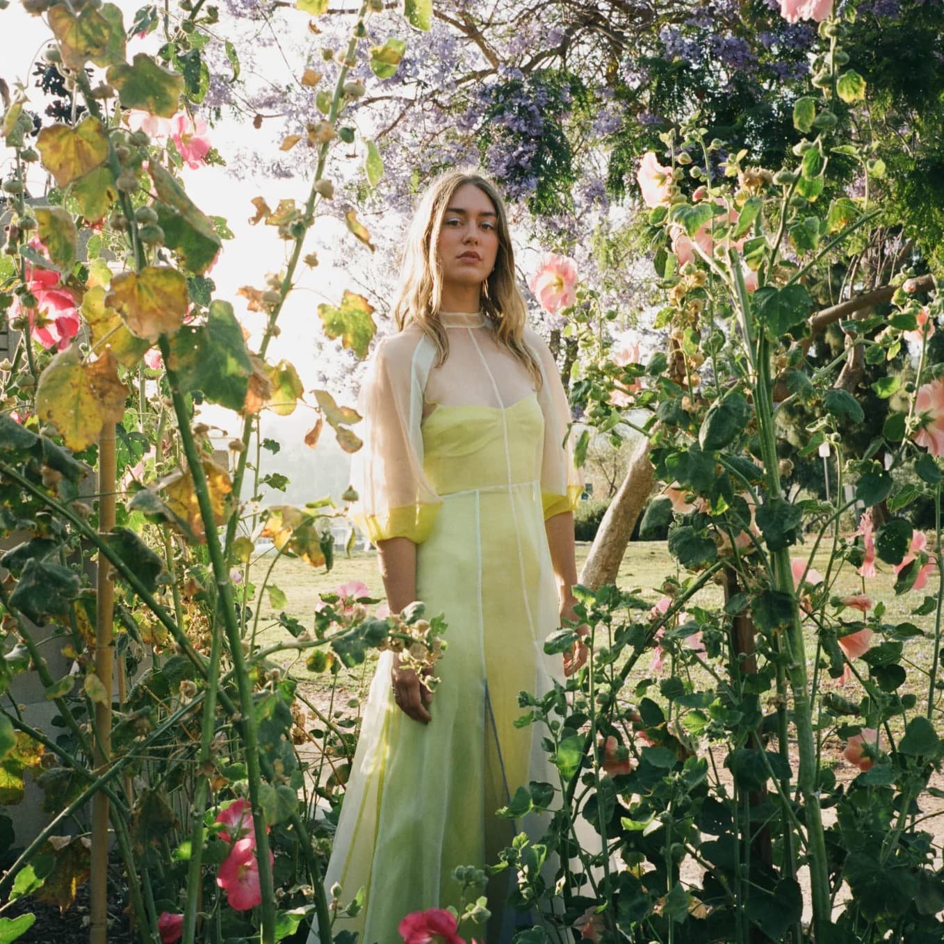 Woman in a pale yellow Kamperett sheer-sleeve dress standing among garden flowers.