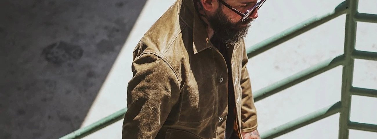 Man in an olive waxed jacket and glasses walking up stairs.